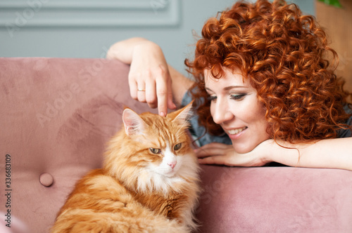 Smiling girl with curly red hair caresses a red cat on an armchair..