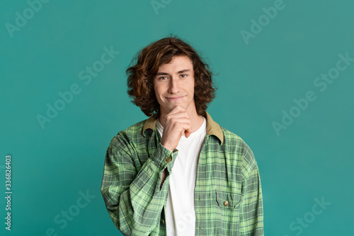 Joyful teenage guy smiling and touching his chin on turquouse background