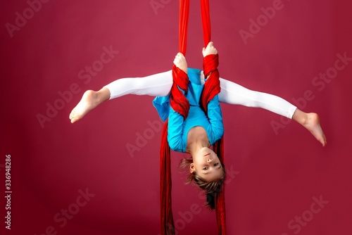 Little acrobat girl doing splits in the air holding on to a red air ribbon on a red-pale background. Acrobatics concept. Advertising space