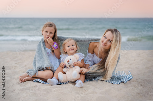 Mom with daughters blondes in white dresses laugh, hug and sit near the blue sea on the beach at sunset and hide behind a gray knitted blanket. Little girl holding a teddy bear.