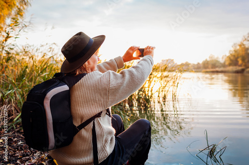 Tourist with backpack taking photos using smartphone of river at sunset. Woman travels admiring spring nature