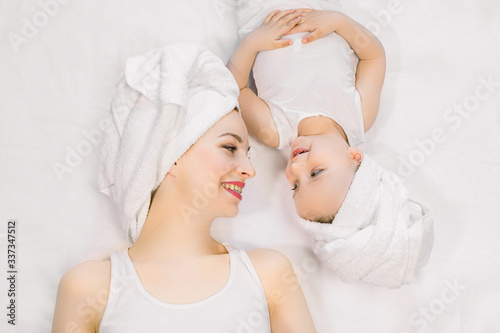 Portrait of happy mother and little teo years old daughter in white t-shirts and towels on heads, lying head to head on the bed and looking each other with smile. Happy family people concept.