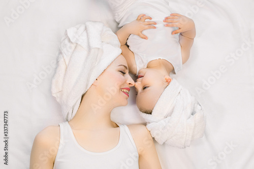 Young pretty relaxed woman mom and her cute little baby girl at home, with towels on heads after bath, lying on bed head to head, looking each other, touching noses and smiling. Happy family spa