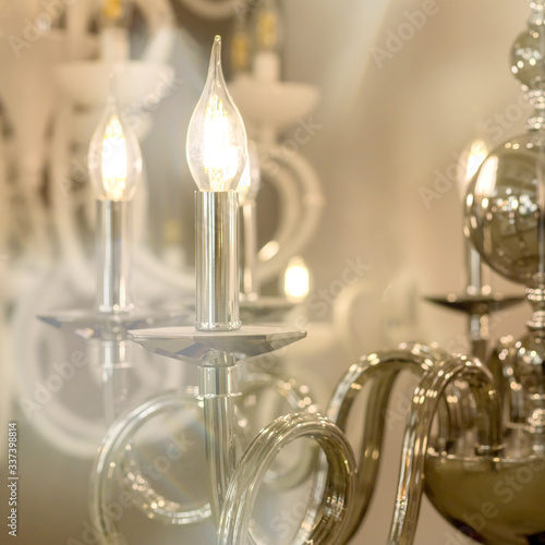 The photography of a glowing chandelier with metal chrome fixtures and shades in the form of candles. The concept of the classical style in the interior. Close up. Soft warm filter, selective focus.