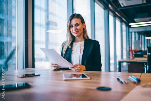 Portrait of successful Caucasian female secretary sitting at desktop with paper documents and looking at camera while working on strategy for corporate organisation of business event with partners