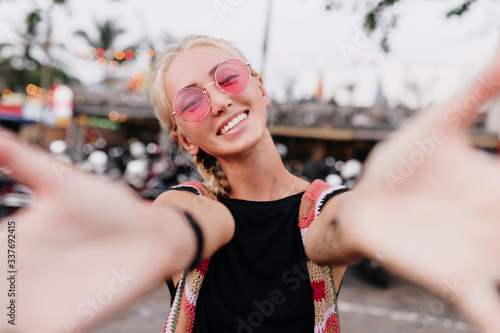 Happy blonde woman with trendy hairstyle fooling around for photo. Laughing tanned lady in pink glasses waving hands on blur street background.