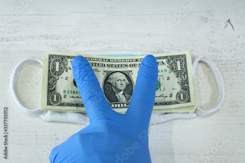 Ludzka ręka w niebieskiej rękawicy ochronnej trzymającej pakiet banknotów i maskę ochronną.