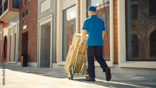Delivery Man Pushes Hand Truck Trolley Full of Cardboard Boxes, Packages For Delivery. Professional Courier Working Efficiently, Quickly. Walks Through Stylish Modern Urban Area. Back View 