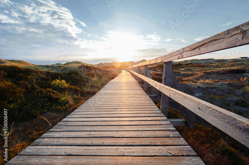 Deserted wooden boardwalk leading away through coastal dunes vegetation towards a glowing cloudy sunset sky.