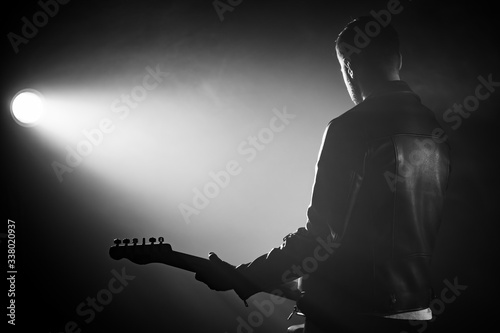 Rock guitarist man in leather jacket standing his back in smoky studio or stage masterfully playing electric guitar. View of unrecognizable musician in the spotlight.