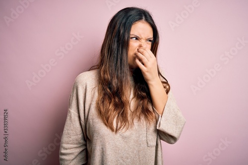 Young beautiful brunette woman wearing casual sweater standing over pink background smelling something stinky and disgusting, intolerable smell, holding breath with fingers on nose. Bad smell
