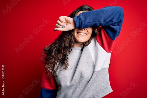 Young beautiful woman with curly hair wearing casual sweatshirt over isolated red background covering eyes with arm smiling cheerful and funny. Blind concept.