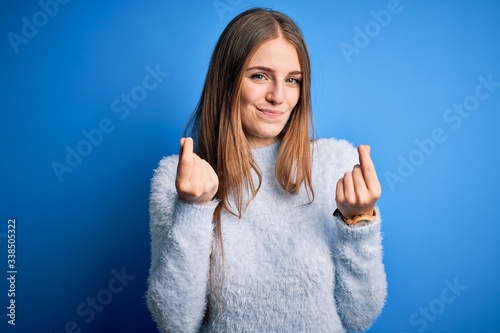 Young beautiful redhead woman wearing casual sweater over isolated blue background doing money gesture with hands, asking for salary payment, millionaire business