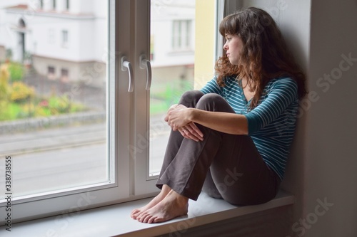 Sad young woman sitting on the window at home isolated, watching out. Coronavirus quarantine concept.