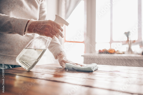 Close up of cleaning home wood table, sanitizing kitchen table surface with disinfectant antibacterial spray bottle, washing surfaces with towel and gloves. COVID-19 prevention sanitizing inside.