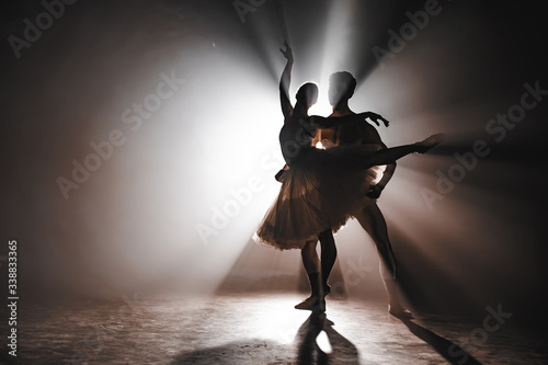 Graceful ballerina and her male partner dancing elements of classical or modern ballet in dark with floodlight backlight. Couple in smoke on black background. Art concept.