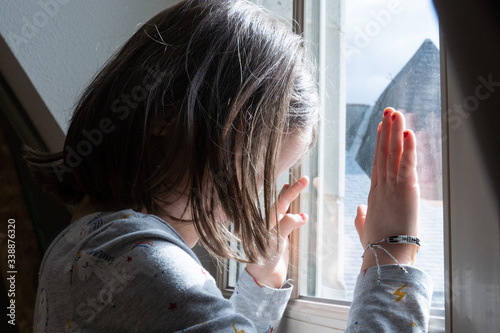 Little girl looking through the window with anxiety during coronavirus quarantine in the house. Stay home concept for families with children. Psychological problems in children in isolation.