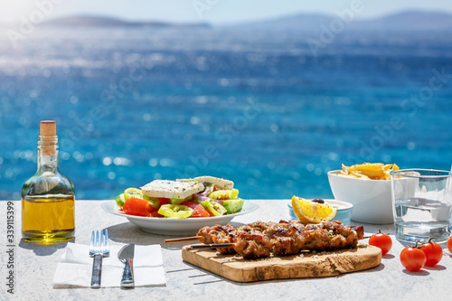 Greek food concept with farmers salad and souvlaki skewers in front of the sparkling, blue Aegean sea during summer time