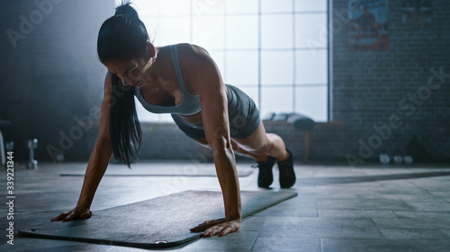 Strong and Fit Athletic Busty Woman in Sport Top and Shorts is Doing Push Up Exercises in a Loft Style Industrial Gym with Motivational Posters. It's Part of Her Cross Fitness Training Workout. 