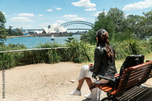 Woman with Sydney Opera House & Harbour Bridge. Tourist looking at attraction, with river water. Blue sky tourism shot. Boats on river. Famous landmark.
