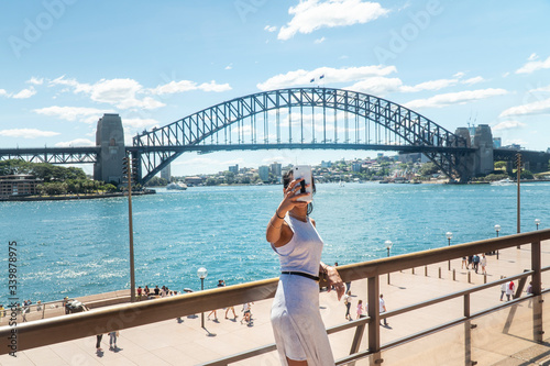Woman taking selfie with mobile phone at iconic Sydney Harbor Bridge. Cityscape, water, with buildings in CBD area. Tourist attraction in Australia. Technology, tourism, travel, taking a photo concept