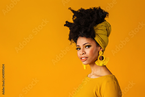 Portrait of a serious young woman with big yellow tassel beaded earrings and afro hair wrapped with head wrap scarf, isolated on yellow background