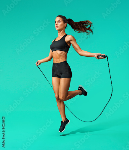 Sporty girl exercising with skipping rope. Photo of girl in black sportswear on turquoise background. Dynamic movement. Full length. Sports and healthy lifestyle