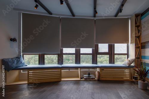 Motorized roller shades in the interior. Automatic roller blinds beige color on big glass windows. Remote Control Shades are above the windosill with pillows. Summer. Green trees outside.