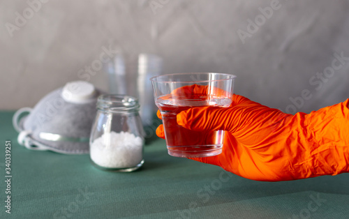 Hand in rubber orange color glove holding plastic cup with salty water to rinse nose and throat as prevention and protection from viruses. Covid-19 concept