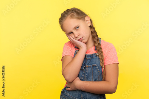 Portrait of unhappy bored little girl with braid in denim overalls almost sleeping on hand and listening to tedious story with disinterest, lazy expression. studio shot isolated on yellow background