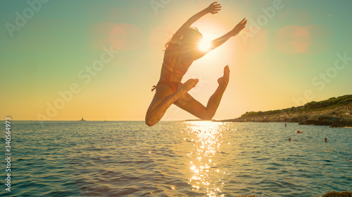 LENS FLARE: Girl jumps off a cliff and kicks legs back while diving into ocean.