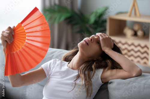 Overheated woman sitting on couch, waving orange paper fan close up, girl feeling unwell, suffering from heating at home, feeling discomfort, hot summer weather or fever, sitting on couch alone