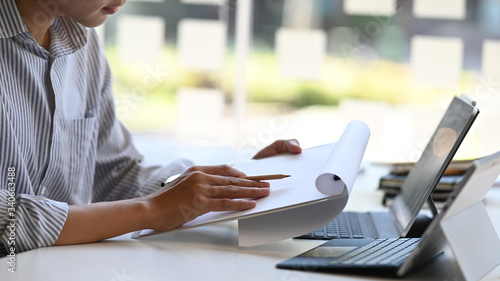 Cropped image of smart man working as architect while planning/drawing on clipboard and sitting at the white working desk with two of computer tablet with keyboard case over glass wall as background.