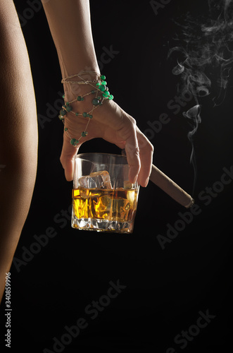 Caucasian model holds glass of whiskey with icecubes and smoking cigar in her hand near naked buttock and hip on dark background closeup studio shot