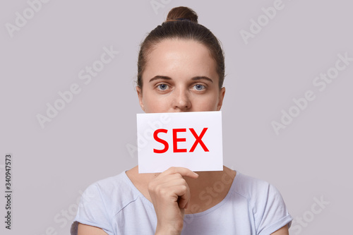 Horizontal image of serious young female with bun looking directly at camera, wearing white t shirt, holding sheet of paper with inscription sex, covering her mouth with paper. Topic concept.