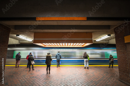 Metro entering fast in a peak transit station with passengers seen from behing standing immobile in front of the speed of the train.