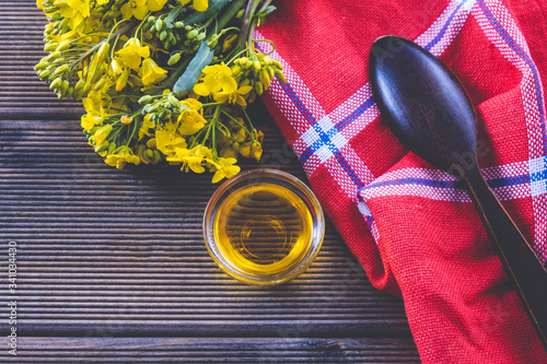 rapeseed flowers near a cup with rapeseed oil and a wooden spoon on a red tablecloth