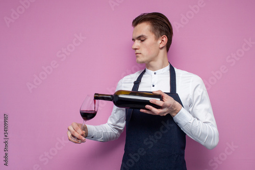 young professional waiter in uniform pours wine into a glass on a pink background, a sommelier guy tasting wine