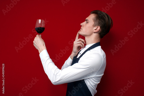 professional thoughtful sommelier holds a glass of red wine and thinks against a red background, the guy the waiter looks at the wine