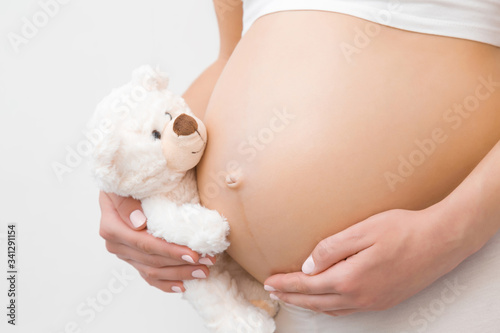 Hand holding smiling white teddy bear. Young woman naked belly. Emotional loving moment in pregnancy time - 30 weeks. Baby expectation. Love, happiness and safety concept. Closeup. Side view.