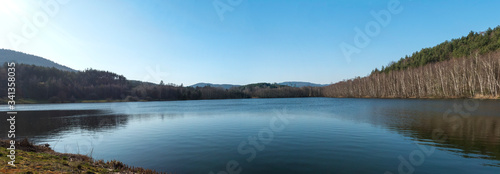 Panoramic view on blue calm water of forest lake, fish pond Kunraticky rybnik with birch and spruce trees growing along the shore and clear blue sky. Nature background. Early spring landscape.