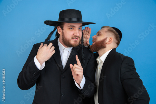 Listening to secrets. Portrait of a young orthodox jewish men isolated on blue studio background. Purim, business, festival, holiday, celebration Pesach or Passover, judaism, religion concept.