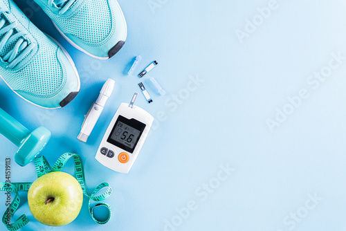 Healthy lifestyle, food and sport concept. Top view of diabetes tester set with athlete's equipment; measuring tape, green dumbbell and fruit on bright blue pastel background.