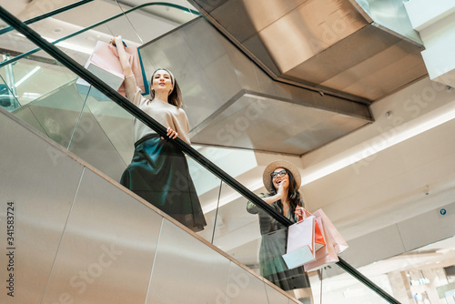 Two girls on an escalator in a shopping center. In the hands of paper bags. Shopping, leisure and entertainment.