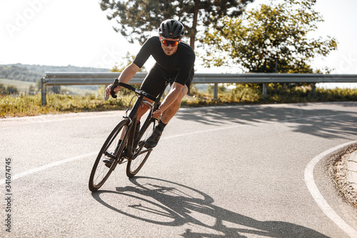 Bearded cyclist in sport clothing, protective helmet and mirrored glasses riding black bike among countryside nature during summer time. Mature man preparing for competitions and races.