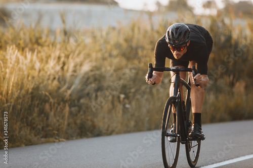 Active professional sportsman cyclist wearing black sports outfit, helmet and glasses riding bike at the paved road outdoors. Strong man improving skills and getting ready for cycling competition