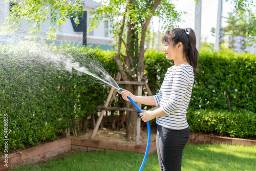 Asian woman having happy in home backyard summer garden, with garden hose splashing water on the lawn and tree leafs during Staying at home using free time about their daily housekeeping routine.