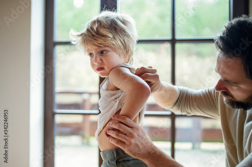 Father with small sick unhappy son indoors at home, checking his back.