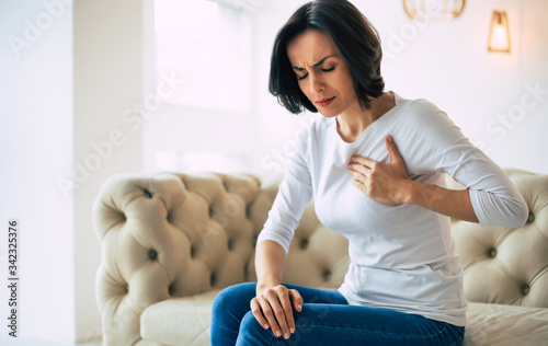 Chest pain. Close-up photo of a brunette girl who is sitting on a couch with her eyes closed and her left hand touching her heart area.