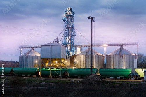 Modern steel agricultural grain granary silos cereal storage warehouse loading railway cargo carriage at night or sunrise. Agribuisness farmland rural industry landscape scene. Mill store facility